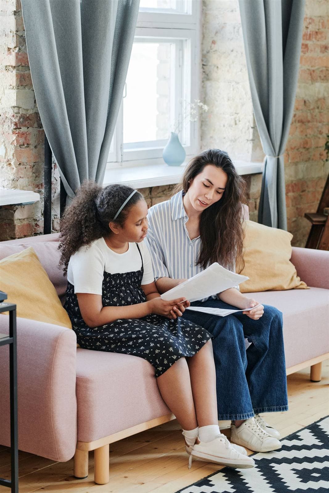 A volunteer and a young girl read homework sheets together on a couch in warm window light