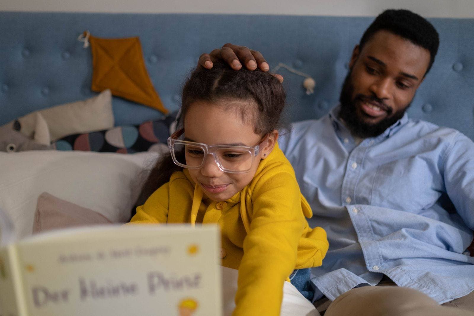 A father smiles and rests his hand on his daughter's head while she leans over a picture book