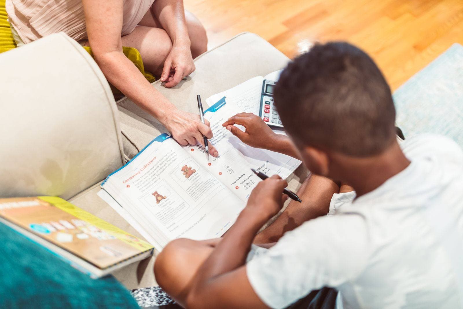 An adult points at a math workbook while a boy works through problems with a pen and calculator